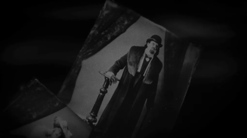 The Role (2013) movie still: closeup — A dark, blurred environment surrounds a handheld black and white photograph. The photo depicts a man in a…