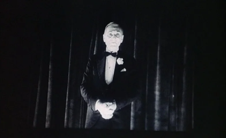 Spirit Of The Beehive (1973) movie still: medium — A man in a tuxedo stands on a dark stage before heavy fabric curtains. The man clasps his hands…