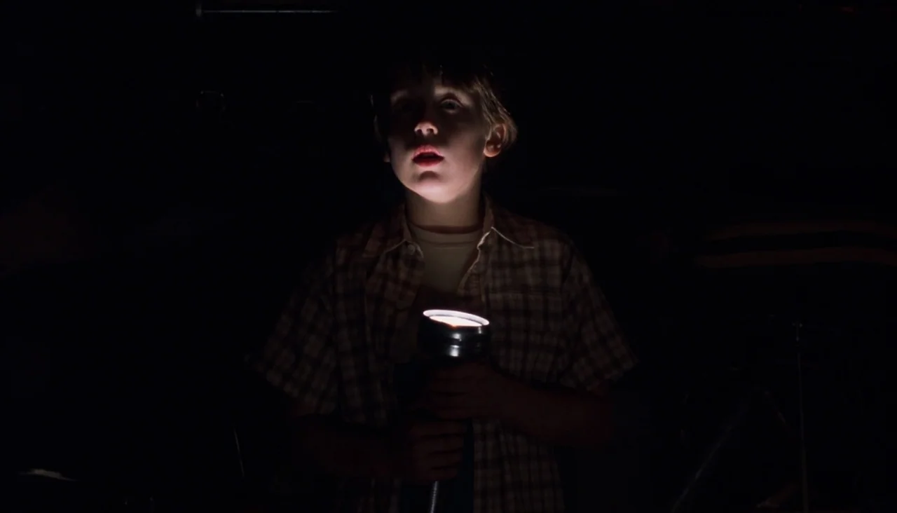 Signs (2002) movie still: medium — The subject stands in complete darkness. He is illuminated only by the light reflecting off his face from a…