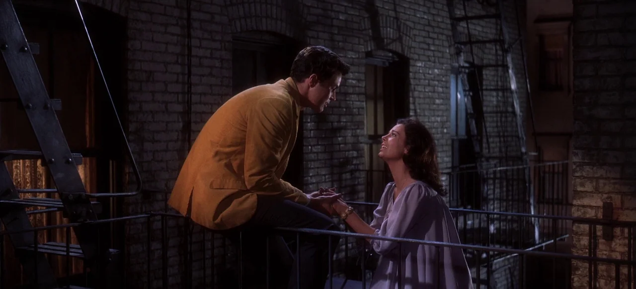 West Side Story (1961) movie still: fullbody — A metal fire escape attached to a dark brick building at night. A young couple sits together on the…