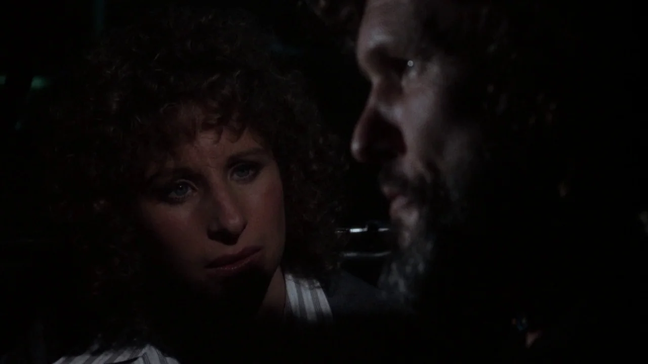 A Star Is Born (1976) movie still: medium — Two individuals sit inside a dark vehicle at night with very minimal lighting. The woman stares at the…