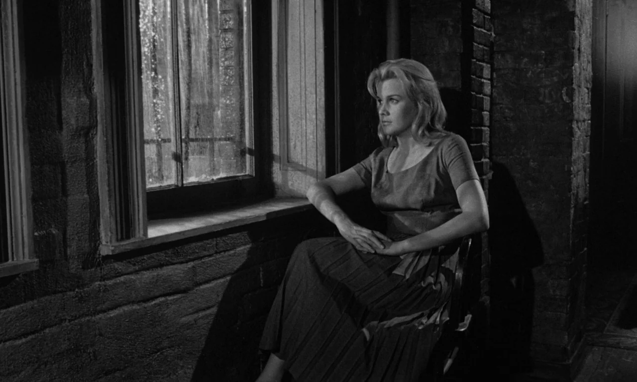 Something Wild (1961) movie still: medium — A woman sits inside near a brick wall by a window at night. She is positioned on a chair while looking…
