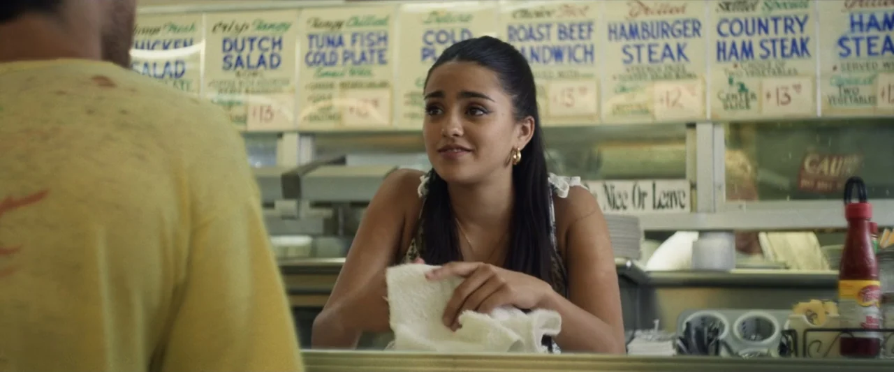 One Fast Move (2024) movie still: medium — A classic diner interior features a menu board posted behind a polished service counter. A young woman in…