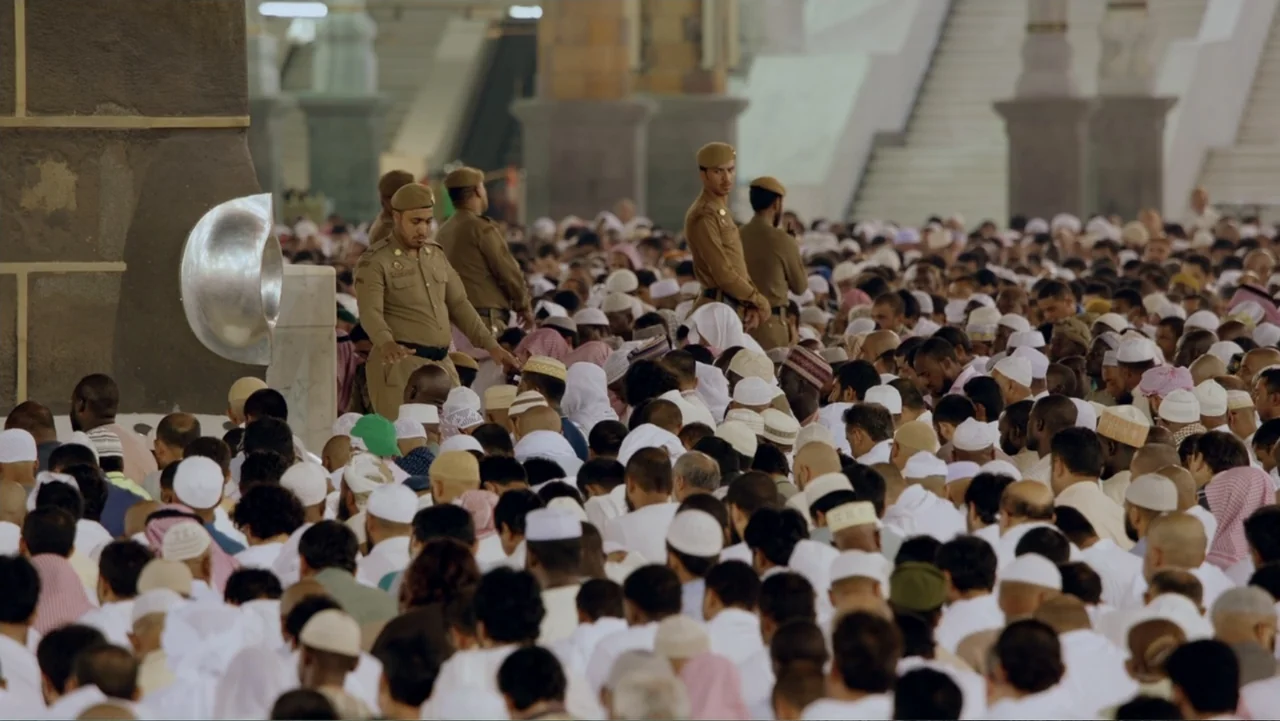 One Day in the Haram (2017) movie still: wide — A crowded interior courtyard features a large assembly of worshippers in white traditional clothing.…