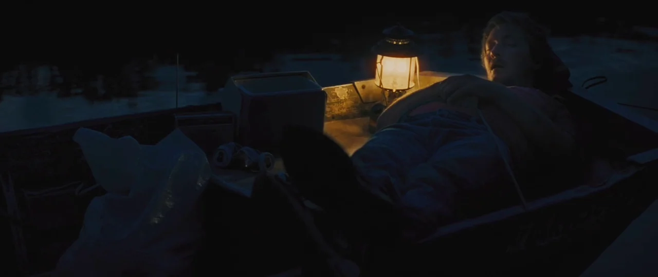 Mud (2012) movie still: medium — A man sleeps inside a small rowboat floating on a dark body of water. A lit metal lantern sits next to him,…