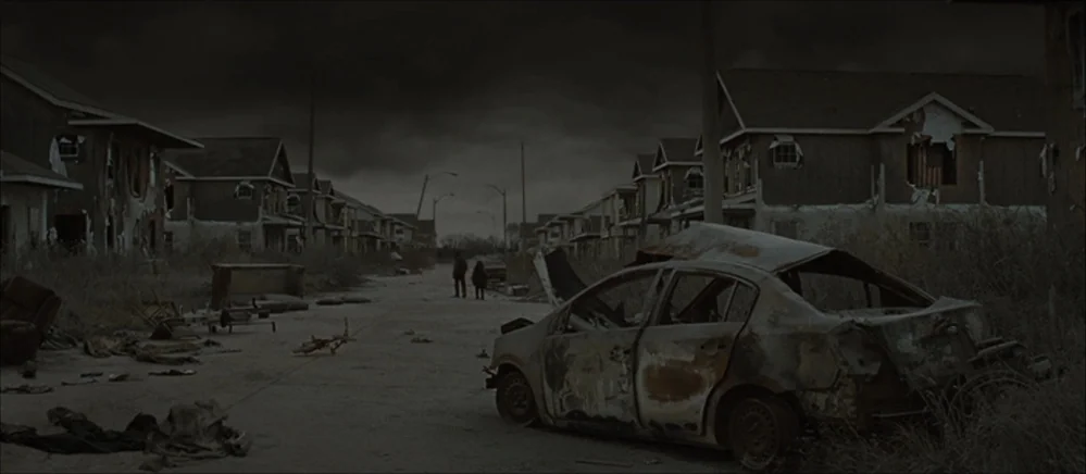 The Road (2009) movie still: wide — A derelict suburban street features rows of decaying houses with peeling facades and debris. A man and a child…