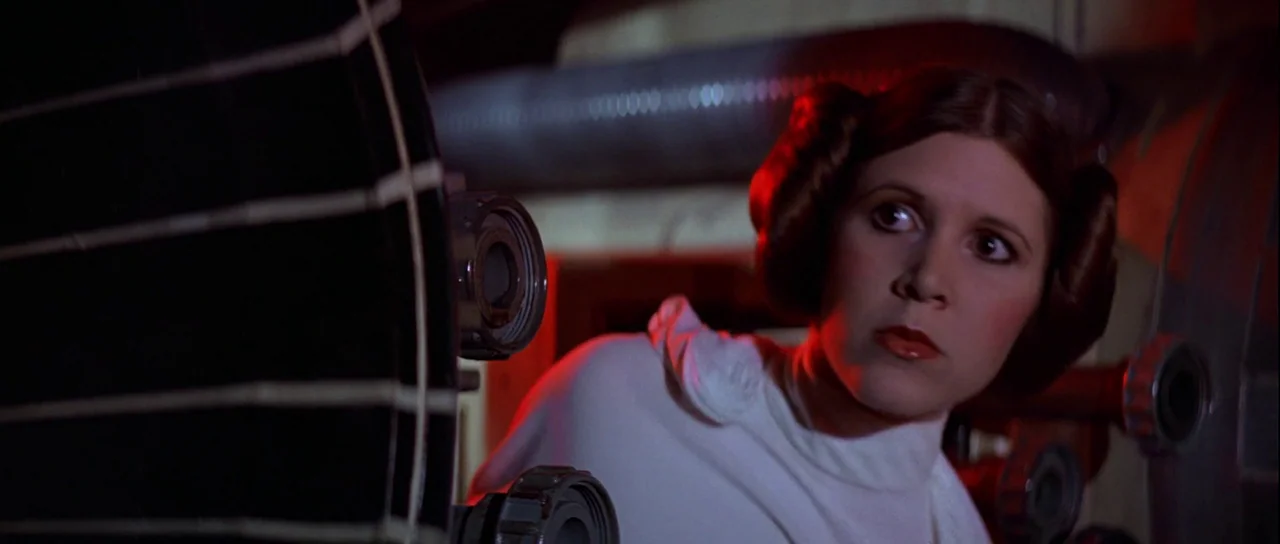 Star Wars (1977) movie still: medium — A dimly lit industrial corridor with metallic pipes and a dark circular structure foregrounded. A woman with…