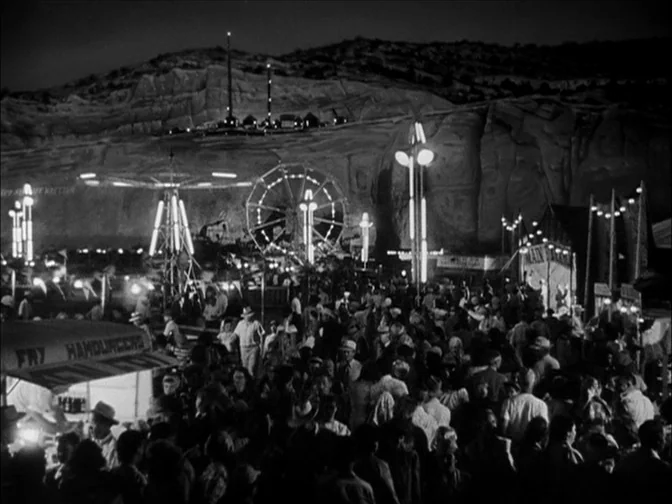 Ace In The Hole (1951) movie still: establishing — A crowded night carnival is nestled against a large, dark, uneven rock formation. A Ferris wheel…