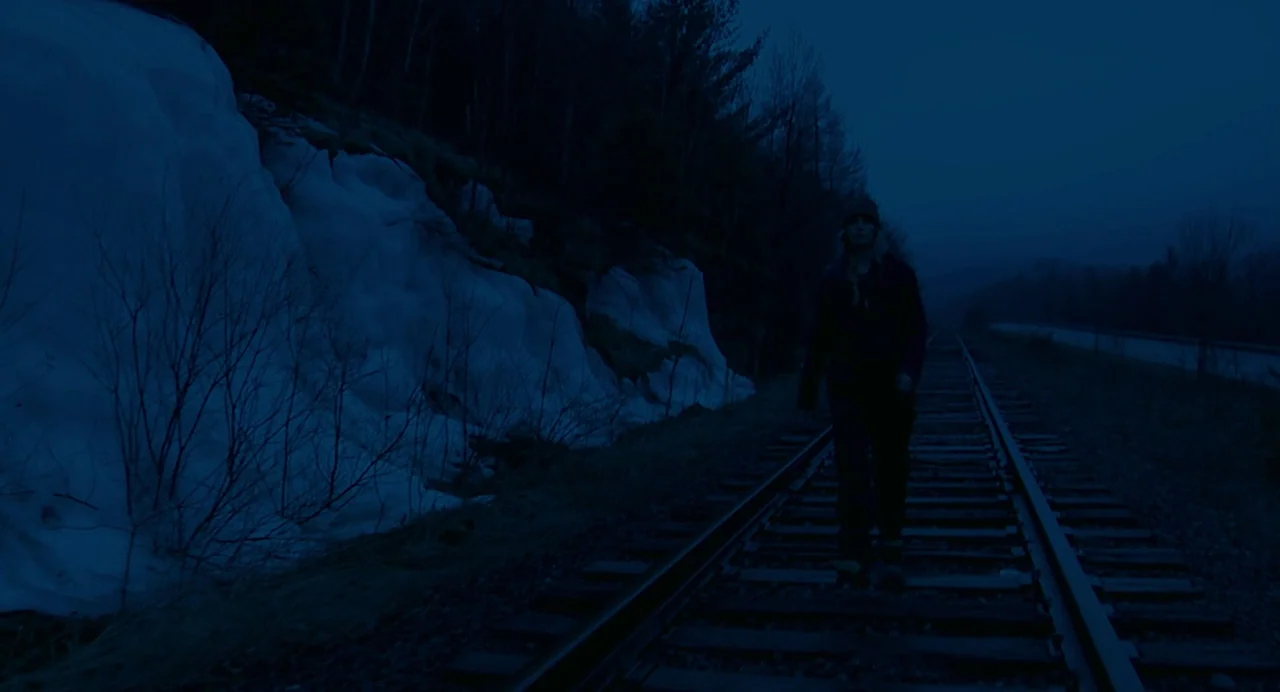 Another Earth (2011) movie still: fullbody — A dark, snow-covered bank sits beside a pair of parallel railroad tracks at night. A young woman walks…