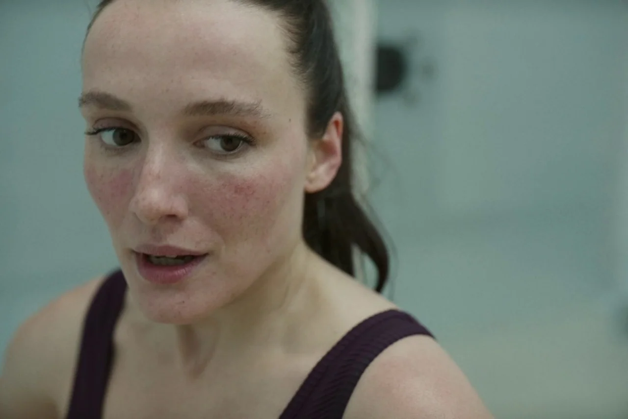 Red Rooms (2023) movie still: closeup — A minimalist bathroom with neutral grey walls and a blurred light-colored background. A woman in a dark…