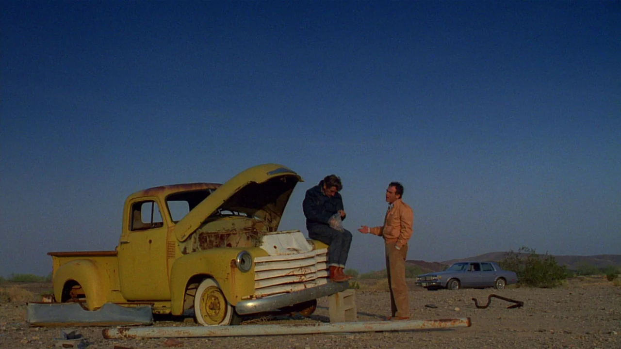 Paris, Texas (1984) movie still: wide — An open, sparse desert landscape under a clear blue sky at sunset. Two men stand beside a rusted yellow…