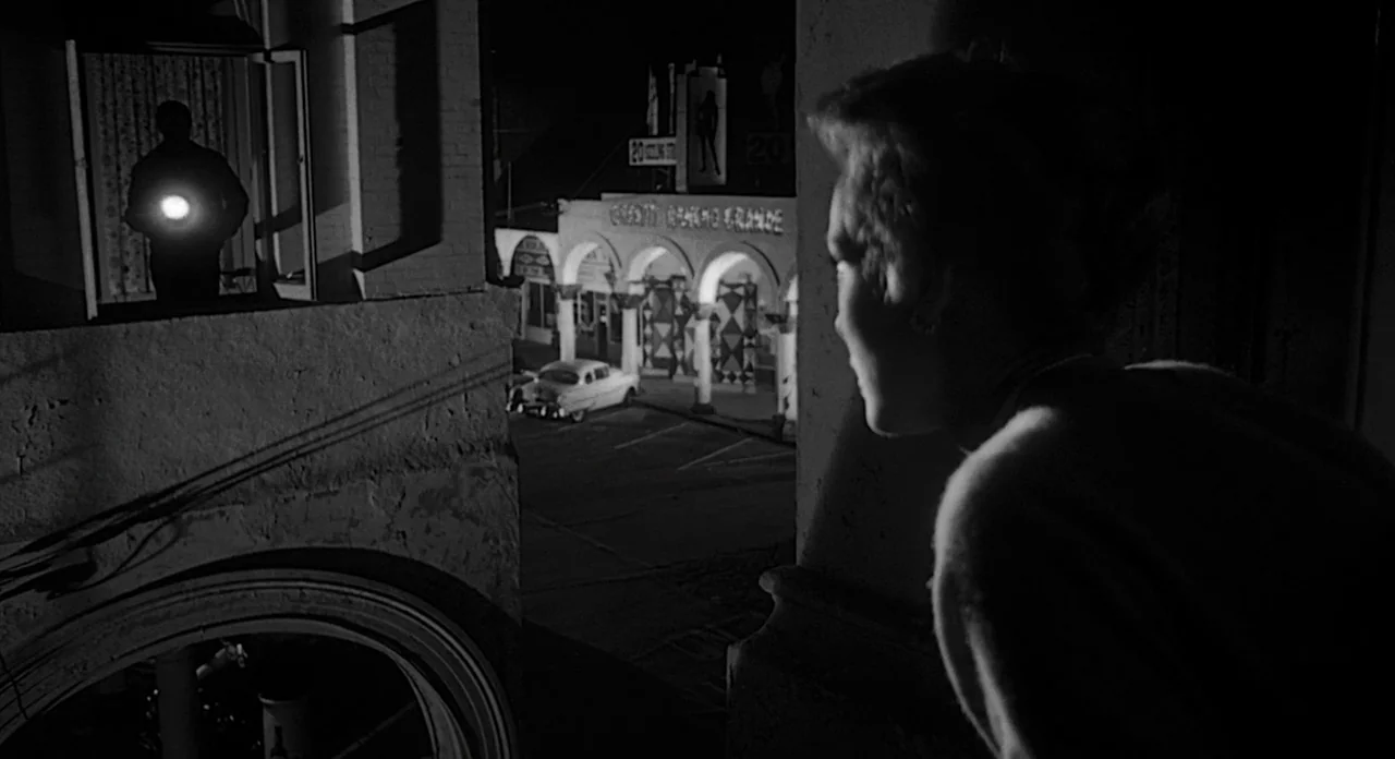 Touch of Evil (1958) movie still: medium — An exterior night view from a balcony overlooking a small town building with arches. A woman stands in…