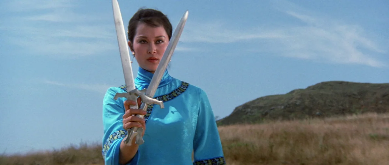 The Legend of the 7 Golden Vampires (1974) movie still: medium — A woman stands in a grassy outdoor field under a clear blue sky. She holds a pair…