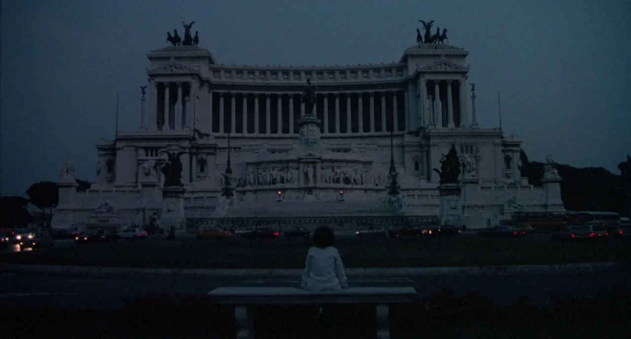 The Belly of an Architect (1987) movie still: wide — A massive white neoclassical monument stands against a dark, twilight sky in Rome. A lone child…
