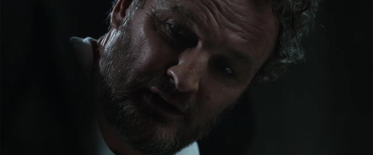 Black Site (2022) movie still: closeup — A dimly lit interior space with a dark, indistinct background. A middle-aged man looks directly toward the…