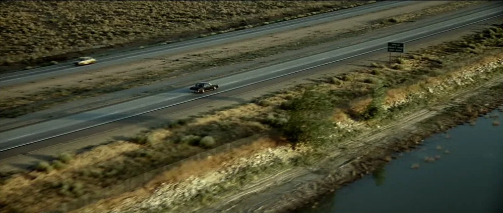 Se7en (1995) movie still: wide — A long stretch of multi-lane highway cuts through a dry and barren desert landscape. A black car drives along the…