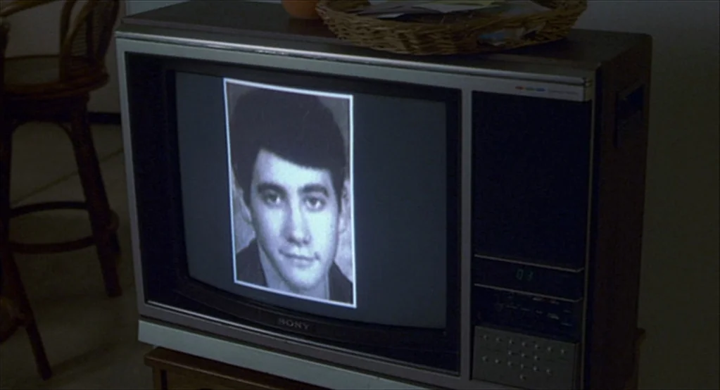 The Good Girl (2002) movie still: medium — A vintage boxy television sits on a wooden stand in a dim room. The screen displays a black-and-white…