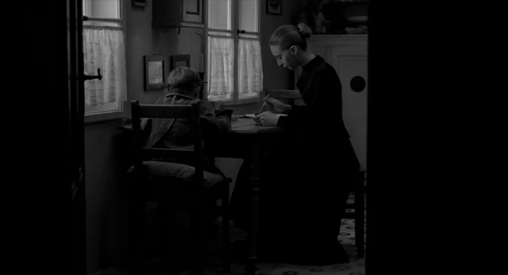 The White Ribbon (2009) movie still: medium — A sparsely furnished room contains a dark wooden table positioned near two windows. A woman in a black…