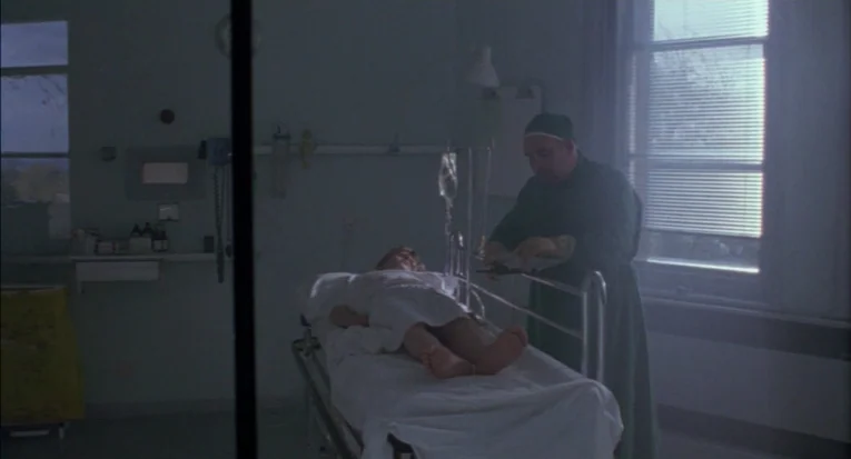 A Room For Romeo Brass (1999) movie still: wide — A sparse hospital room contains a gurney with a patient and medical equipment. A doctor in scrubs…