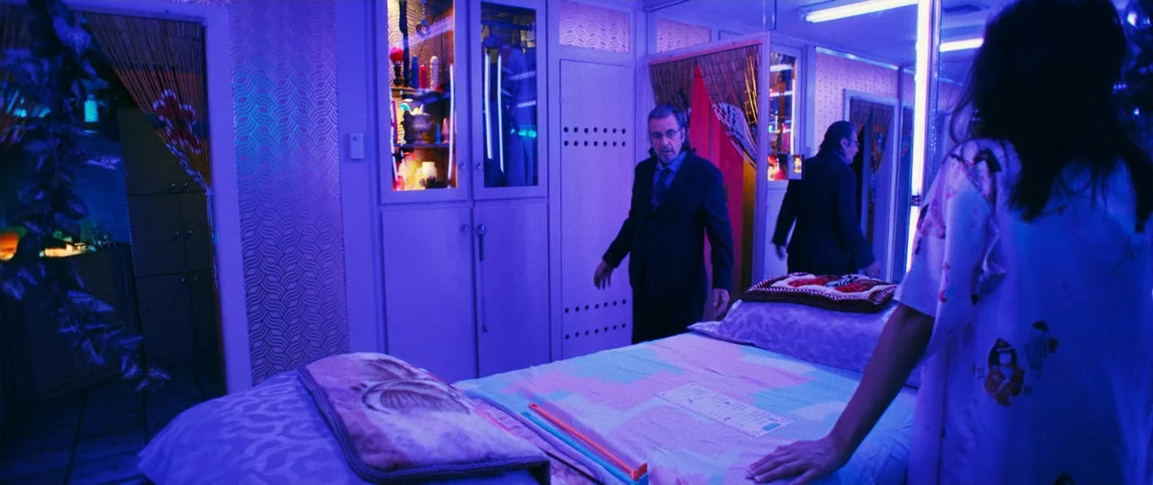 Manglehorn (2014) movie still: medium — A small room with neon lighting and patterned walls features a massage table centerpiece. A man in a suit…