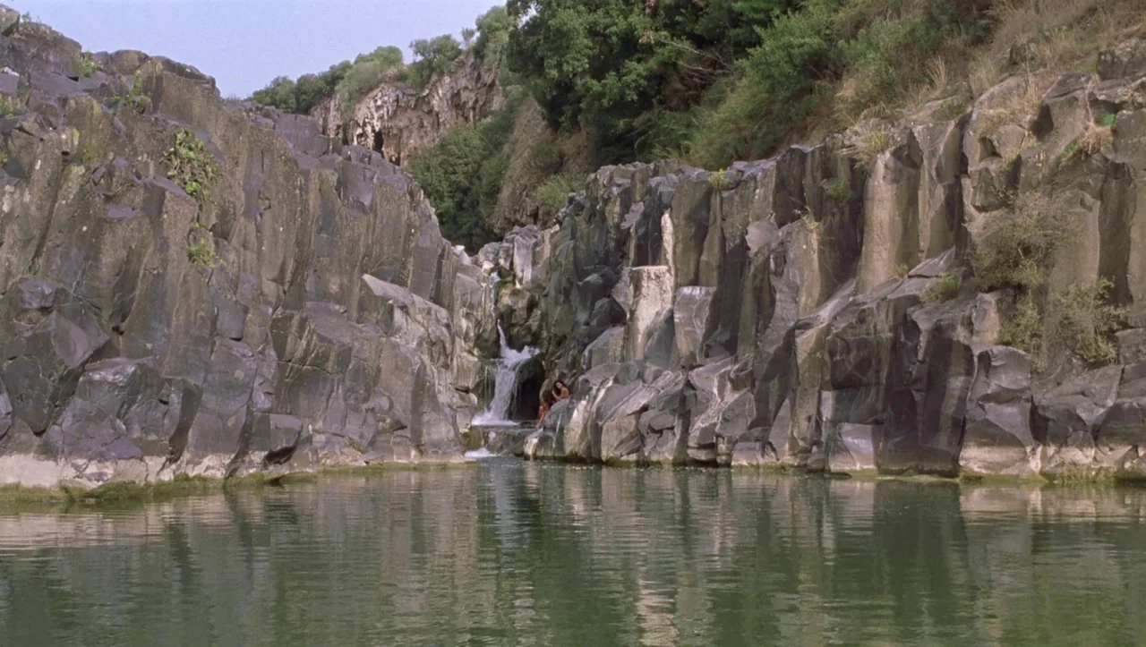Eros (2004) movie still: wide — A narrow pool of water is framed by tall, jagged basalt canyon walls. Two people sit quietly on a rock ledge near a…