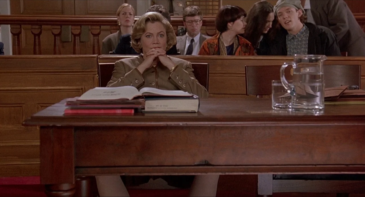 Serial Mom (1994) movie still: medium — A wood-paneled courtroom features a long desk in the foreground and gallery seating behind. A woman sits at…