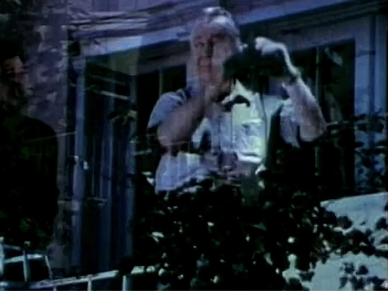 In the Year 2889 (1969) movie still: medium — An exterior balcony at night features dark foliage and a glimpse of an interior window. A man in a…