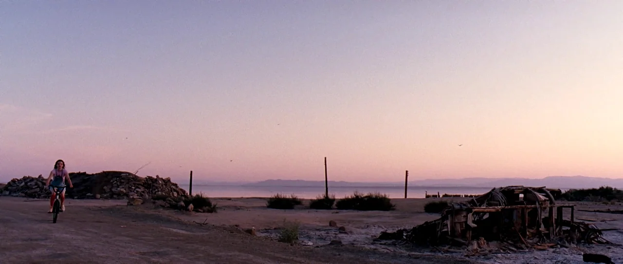 Little Birds (2011) movie still: wide — A desolate shoreline landscape features piles of rubble and the frame of a wrecked vehicle. A lone teenage…