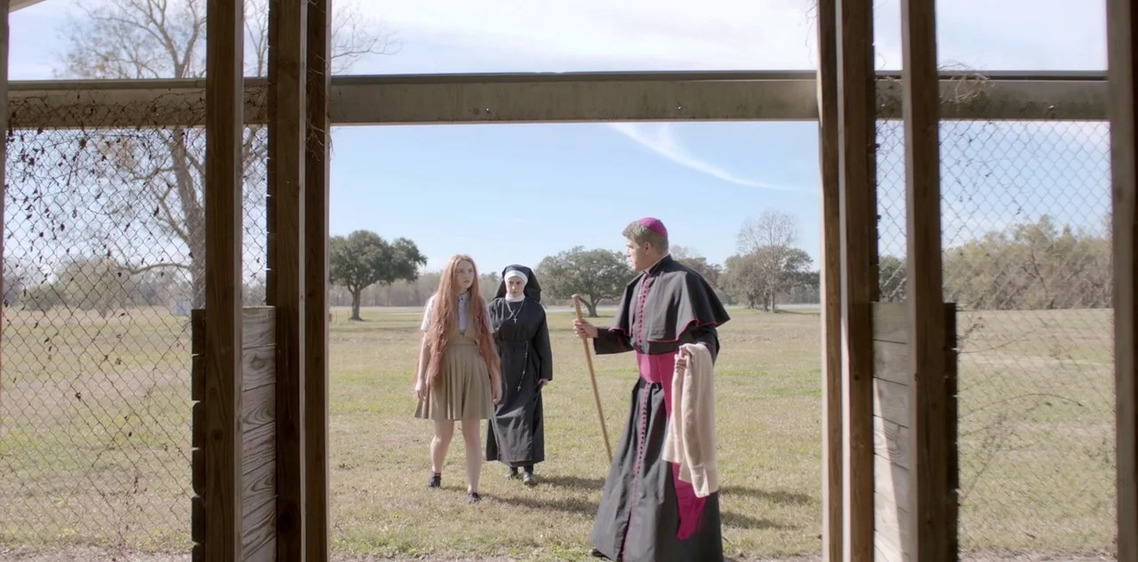 Darlin’ (2019) movie still: fullbody — A flat rural landscape stretches out behind a wooden fence structure during the day. A bishop carrying a…
