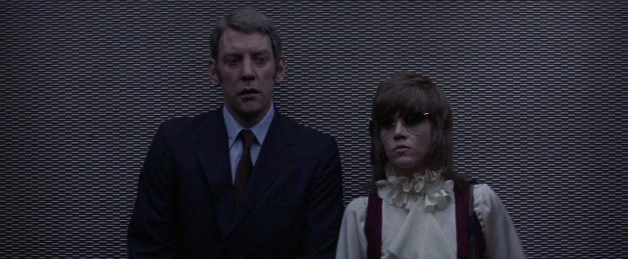 Klute (1971) movie still: medium — Two people stand inside an elevator with gray textured metallic walls behind them. A man in a dark suit stands…