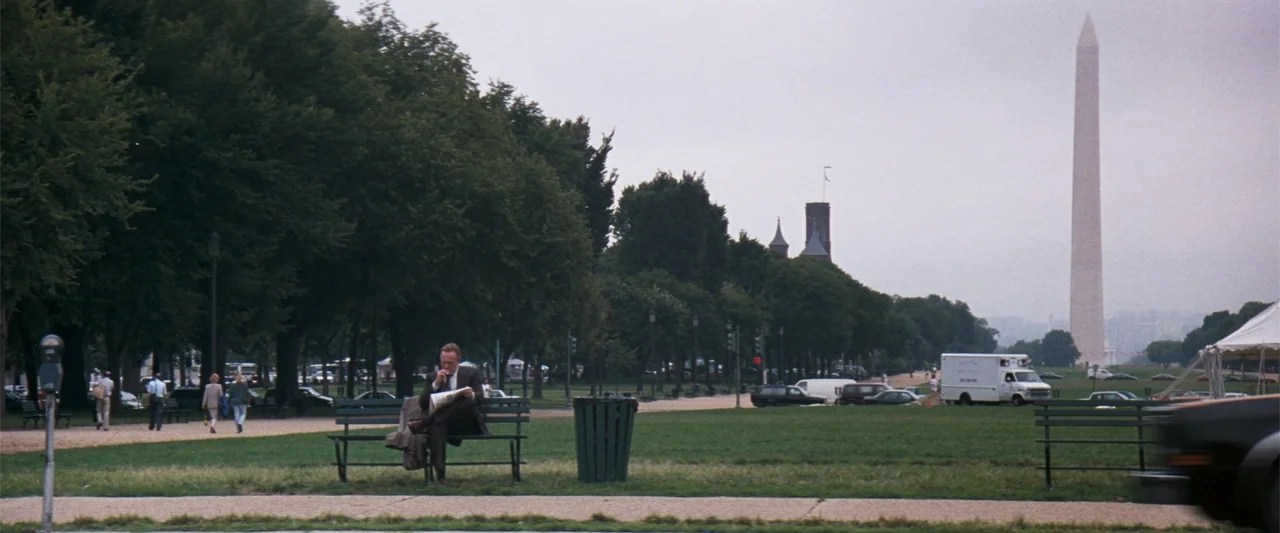 The Eraser (1977) movie still: wide — A spacious green park features rows of mature trees and the Washington Monument afar. A man sits alone on a…