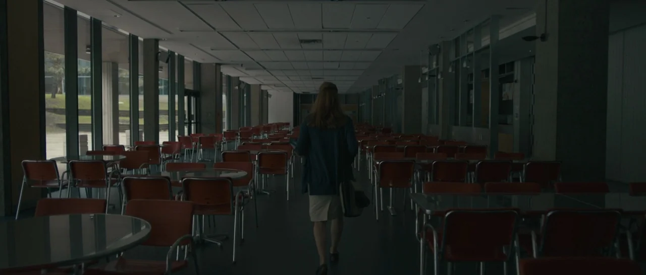 Arrival (2016) movie still: fullbody — A spacious, sparsely populated modern cafeteria features rows of red plastic chairs and tables. A woman walks…