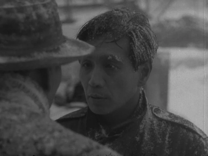 The Idiot (1951) movie still: medium — An outdoor winter setting with falling snow visible on the subjects' clothing and hair. A man looks at an…