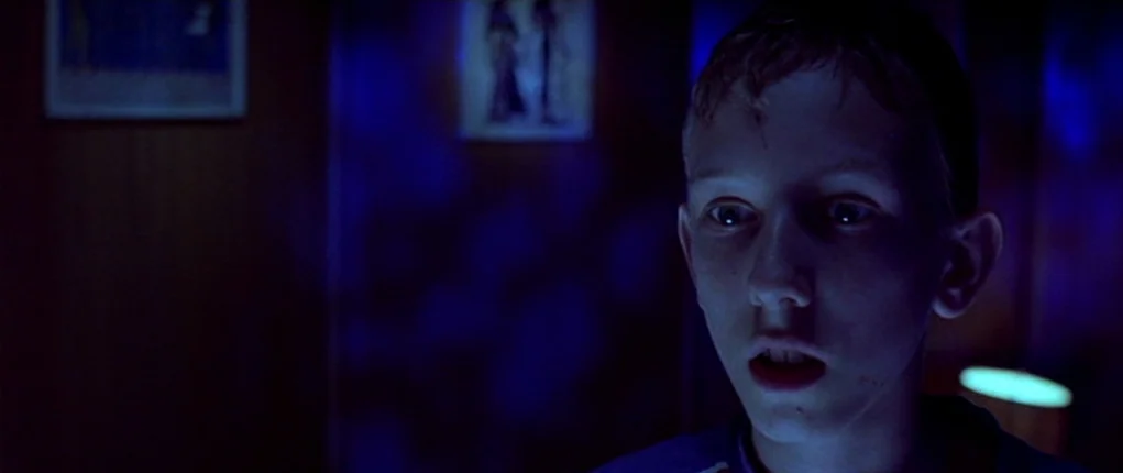 I Want You (1998) movie still: closeup — A dimly lit room with blue ambient light originating from a screen. A young boy stands facing the light…
