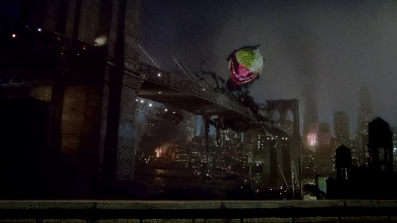 Little Shop of Horrors (1986) movie still: wide — A massive plant monster looms over the Brooklyn Bridge against a dark, foggy night cityscape. The…