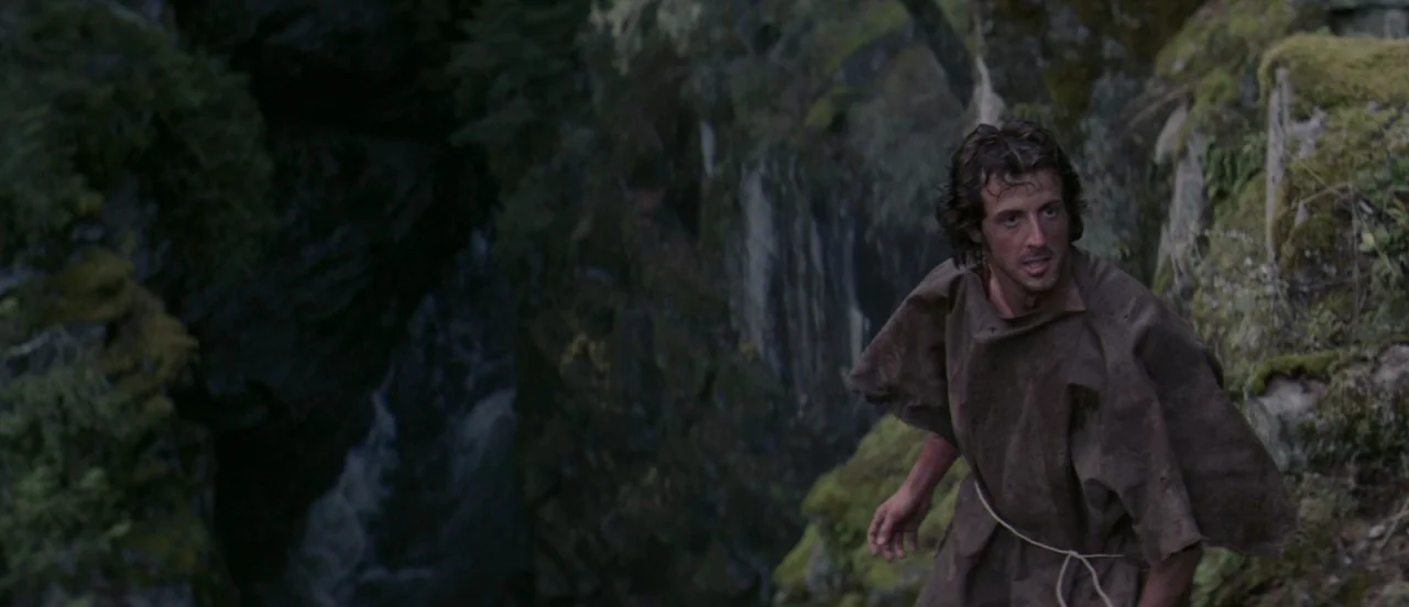 First Blood (1982) movie still: medium — A dark rocky canyon wall is covered in green moss and shadows. A man in a ragged poncho climbs cautiously…