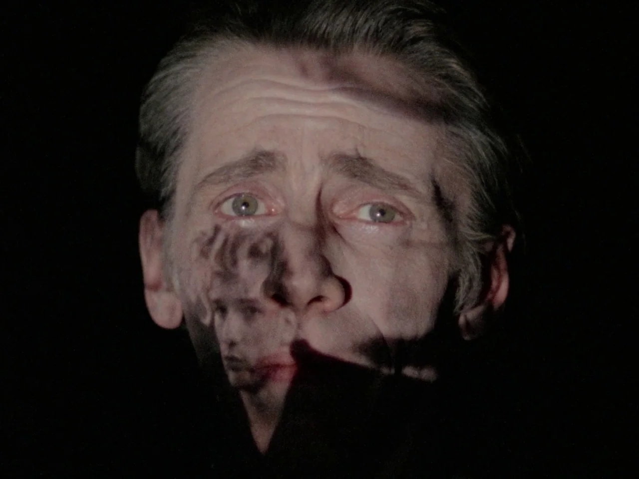Decoder (1984) movie still: closeup — A man's face is illuminated against a solid black background. An image of another person is projected onto his…