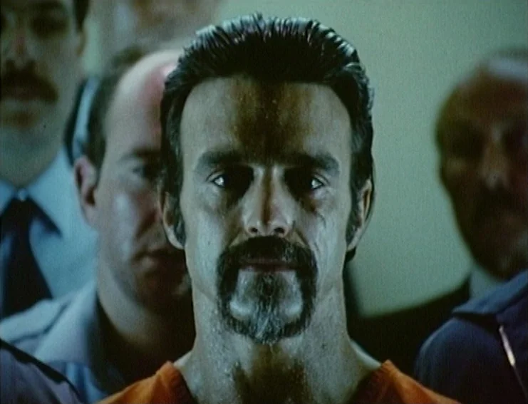 Ghosts… Of The Civil Dead (1988) movie still: closeup — A man stands in a dimly lit, nondescript room surrounded by other figures. He stares…