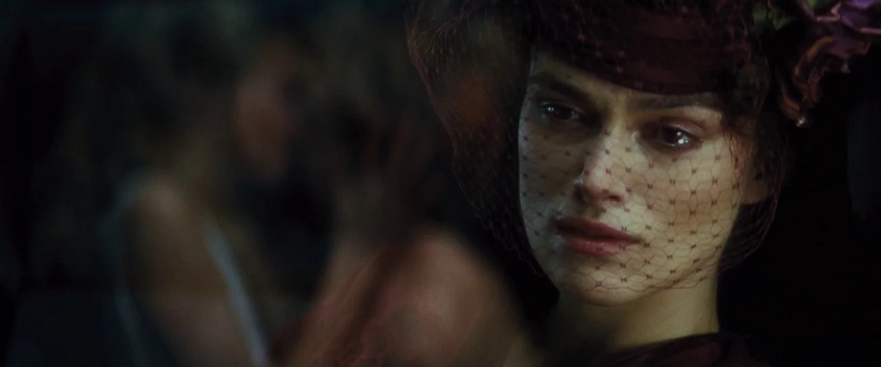 Anna Karenina (2012) movie still: closeup — A dark carriage interior with a woman wearing a decorative red veiled hat. She sits in partial shadow…