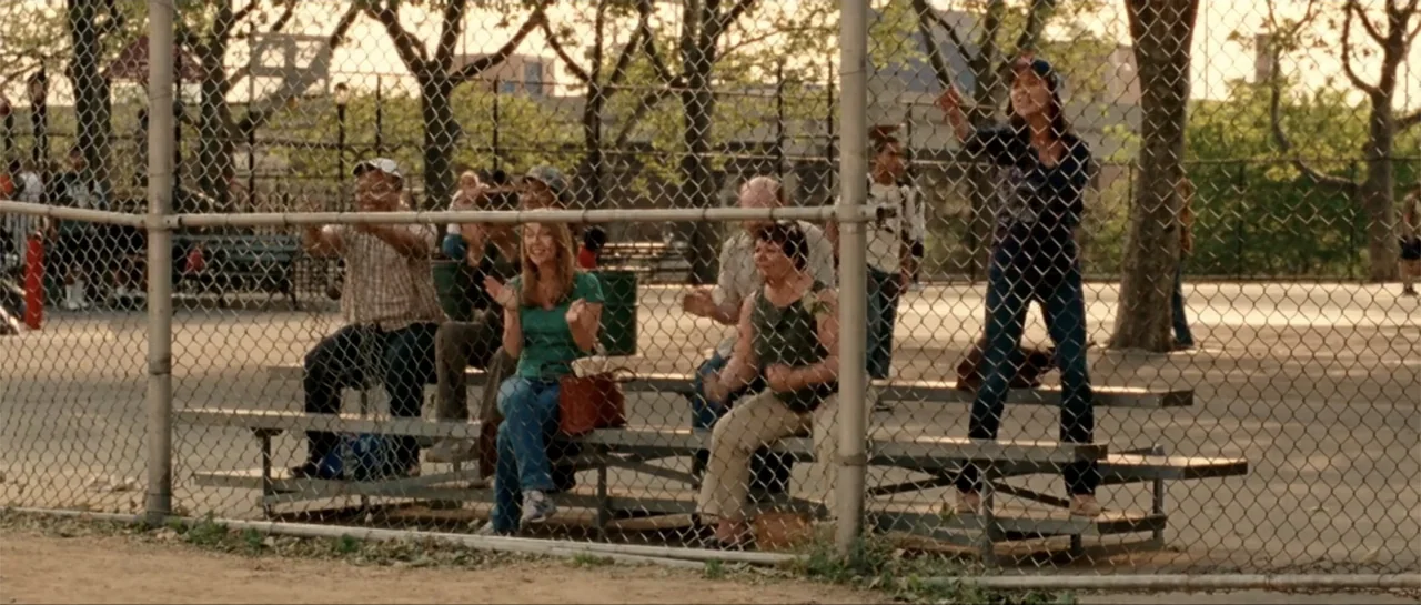 The Rebound (2009) movie still: medium — A group of spectators sits on metal bleachers behind a baseball field chain link fence. The people are…