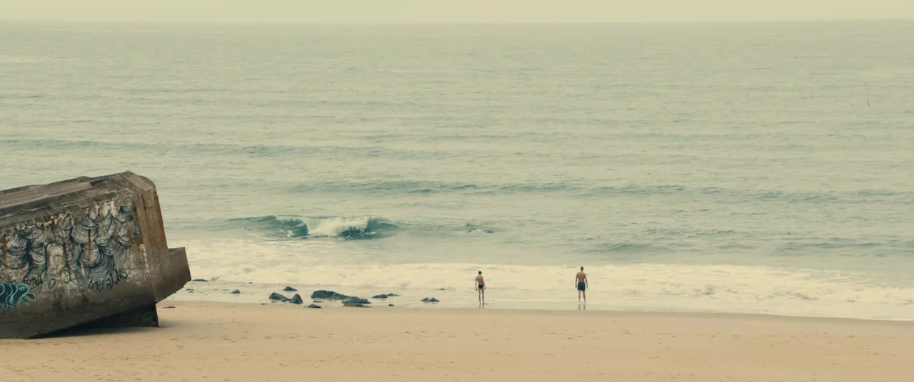 BPM (Beats Per Minute) (2017) movie still: wide — A wide sandy beach features a weathered concrete bunker with dark blue graffiti drawings. Two men…