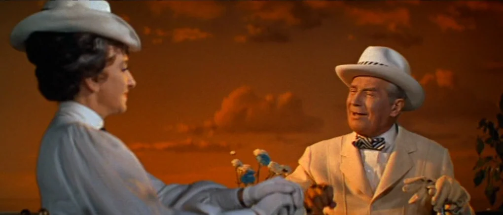 Gigi (1958) movie still: medium — An outdoor balcony at sunset features a painted sky backdrop of orange clouds. A man and woman in formal white…
