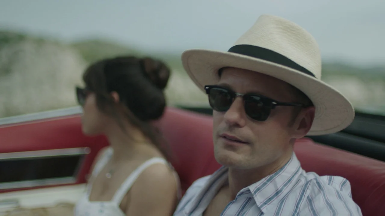 Infinity Pool (2023) movie still: medium — A man and woman sit in a red-upholstered convertible vehicle during the day. The man looks toward the…