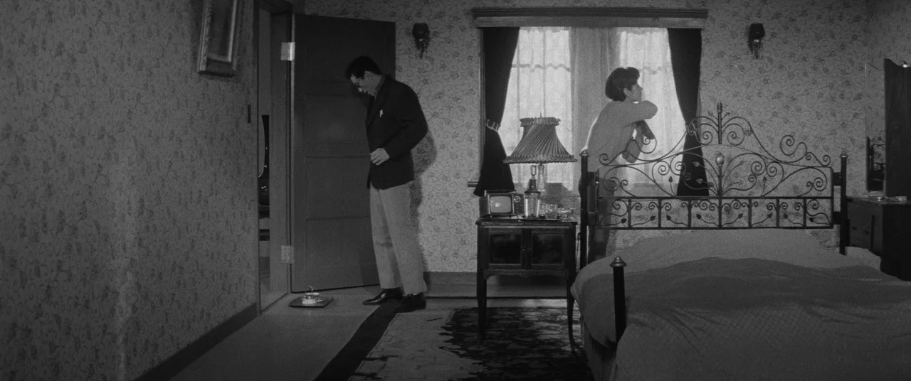 Carmen From Kawachi (1966) movie still: wide — A hotel room features floral patterned wallpaper, an iron bed frame, and sheer curtains. A man stands…