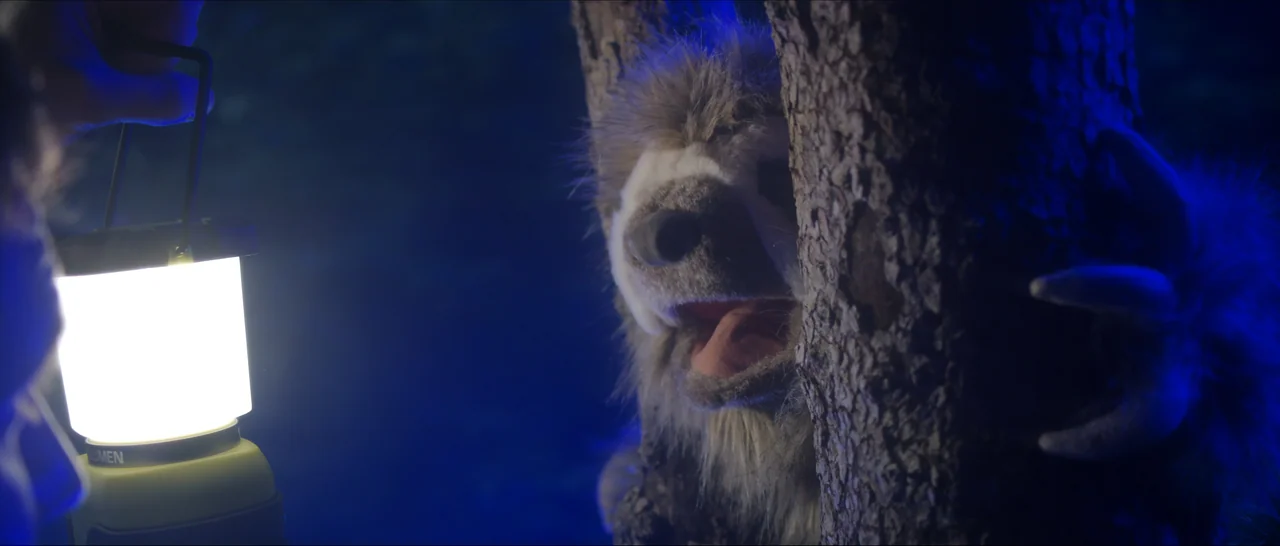 Songs For A Sloth (2021) movie still: medium — A dark, blue-lit outdoor setting at night features a large, rough tree trunk. A furry creature peeks…