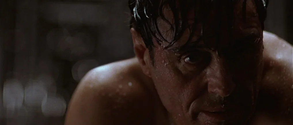 Sexy Beast (2000) movie still: closeup — A man stands in a dark environment with water beading on his skin. His face is illuminated against deep…