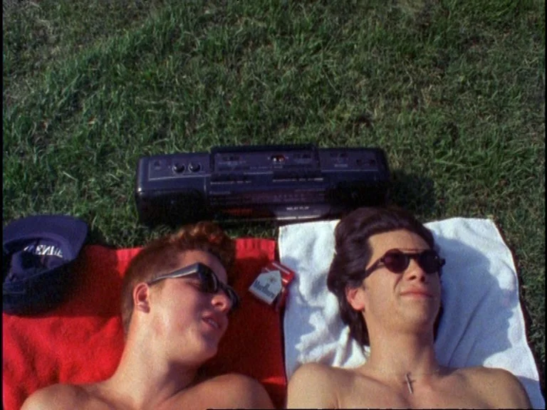 Totally F***ed Up (1993) movie still: medium — Two men lie on red and white towels spread out over a green lawn. A boombox and a pack of cigarettes…