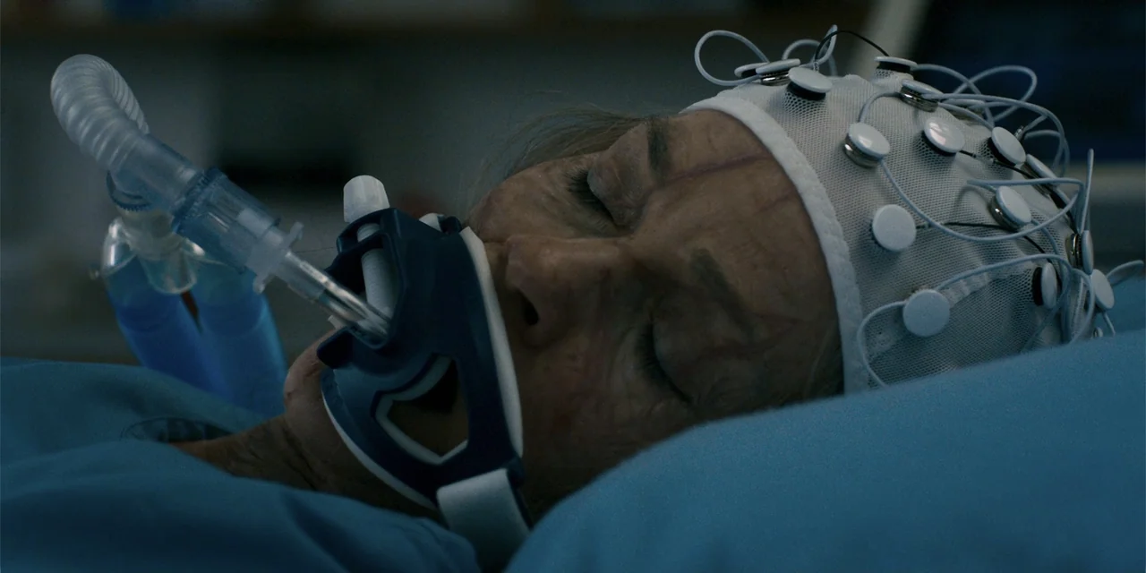 Demonic (2015) movie still: closeup — A sterile medical environment features a person resting under monitoring equipment. An elderly woman lies in a…