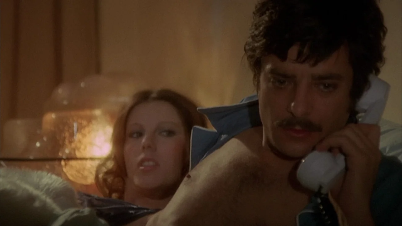 The Black Belly of the Tarantula (1971) movie still: medium — A dimly lit room at night with warm background lighting highlighting the figures. A…