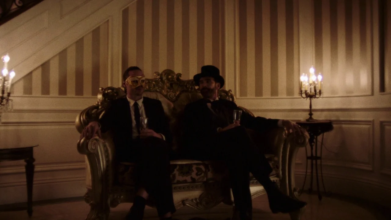 Snowbound (2017) movie still: medium — An opulent room with beige striped wallpaper and ornate gold molding contains a decorative sofa. Two men in…